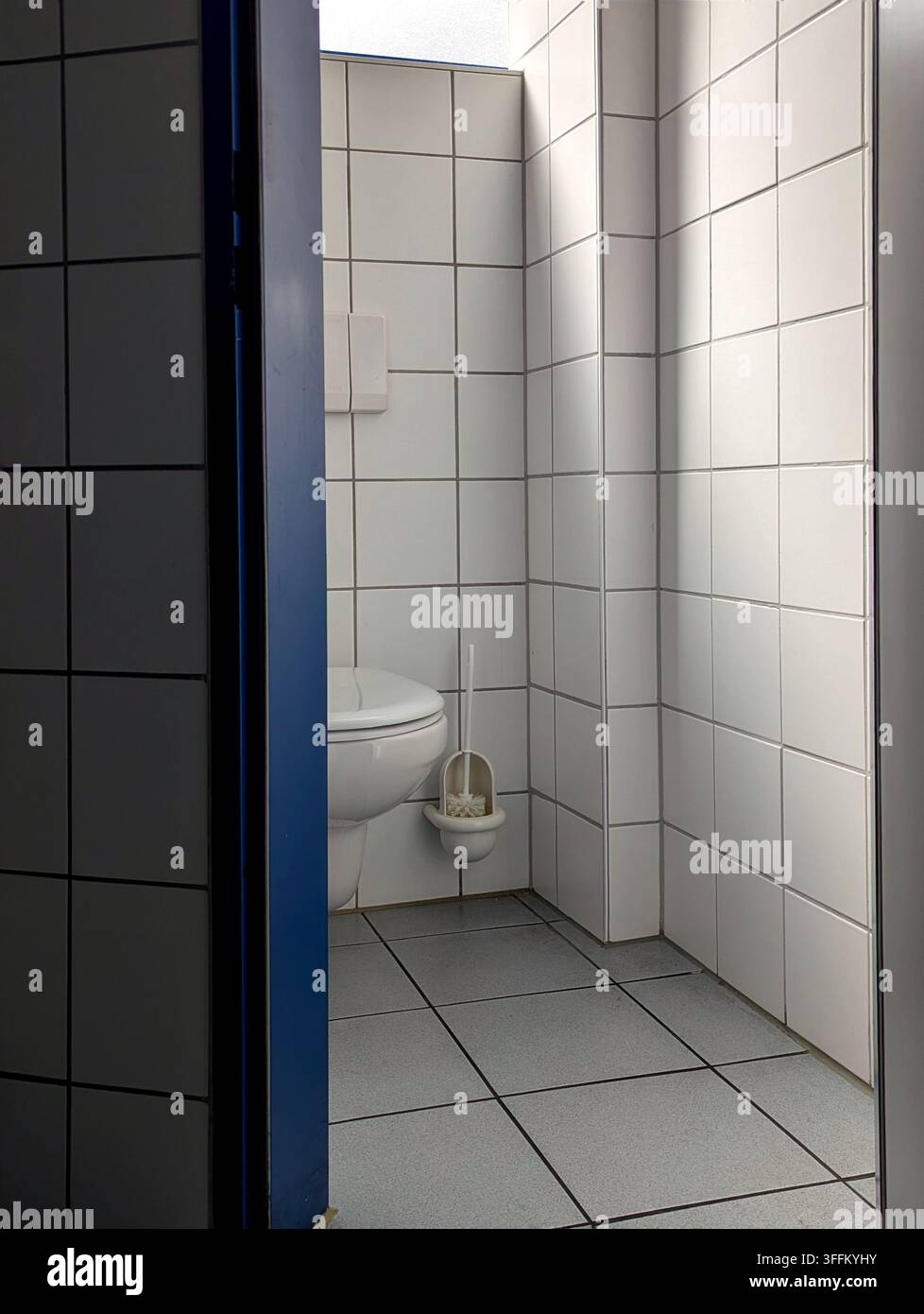 Una foto chiara e pulita che guarda attraverso una cornice di porta blu in una cabina di servizi igienici pubblici. La cabina presenta piastrelle murali quadrate bianche, piastrelle del pavimento grigie Foto Stock