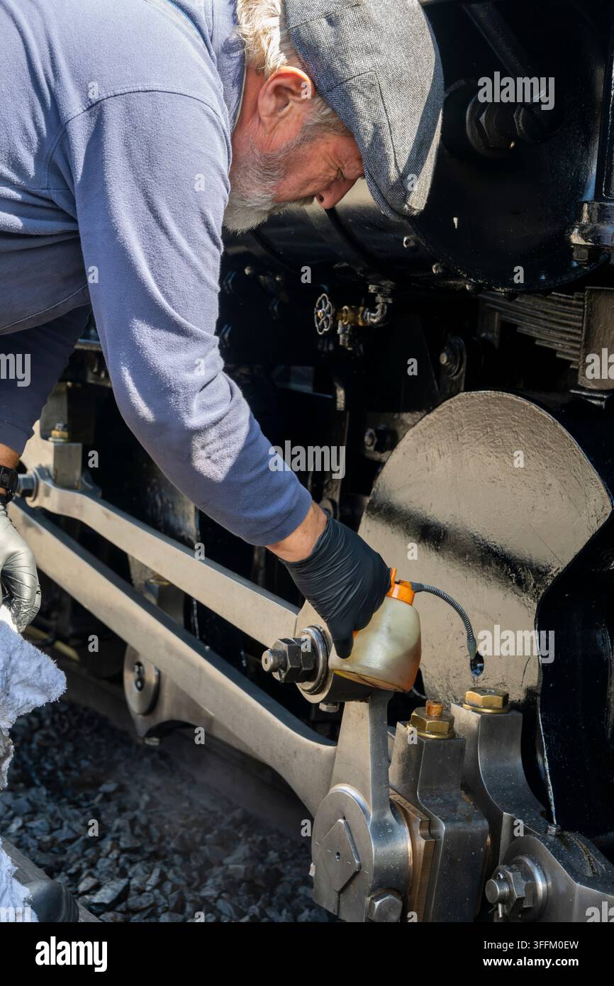 Un uomo con un berretto piatto e guanti applica olio alla complessa meccanica di una storica macchina a vapore Foto Stock