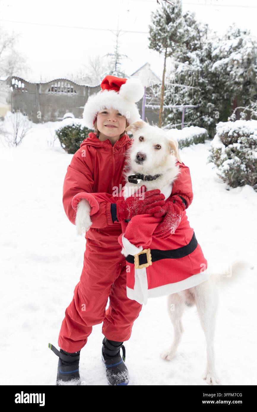 Ragazzo in un pigiama da neve rosso abbraccia il suo cane vestito con una giacca di Babbo Natale mentre cammina in un parco coperto di neve Foto Stock