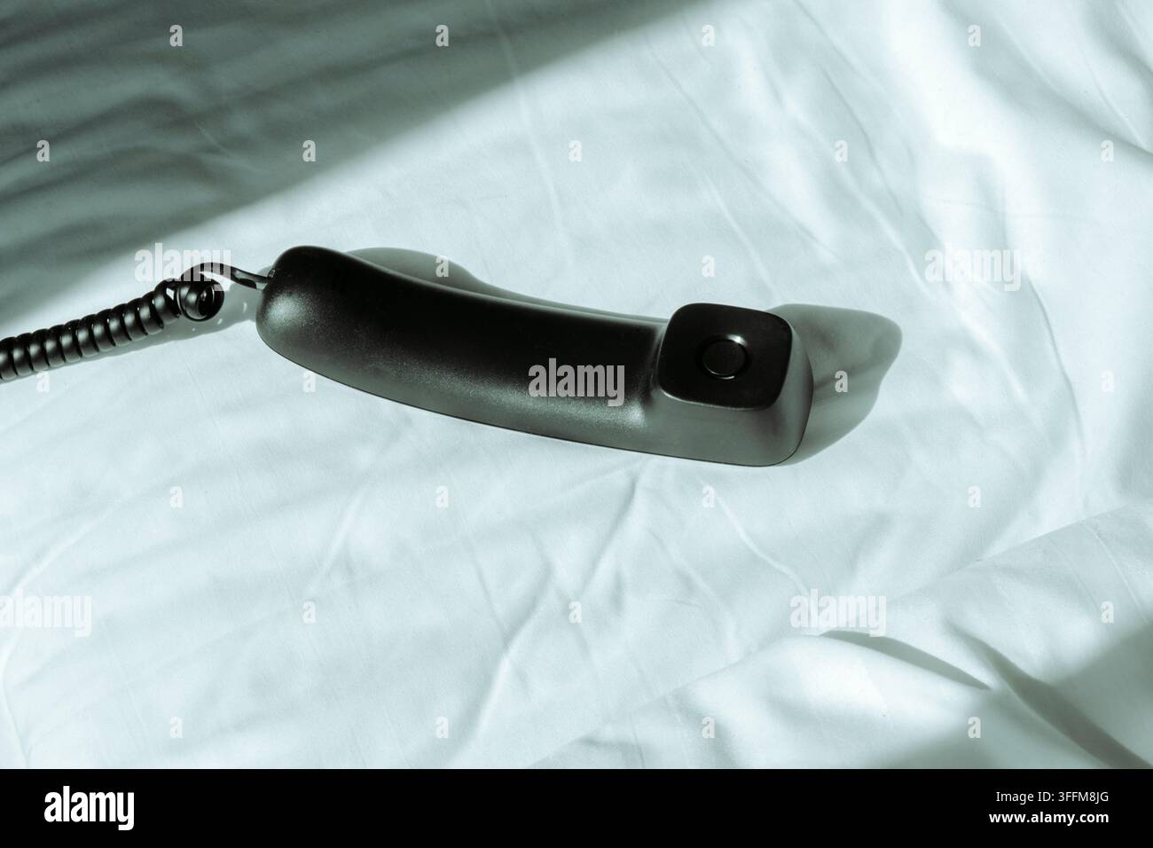 Un auricolare nero di telefono fisso con cavo avvolto giace sulle lenzuola bianche sgualcite di un letto disfatto, proiettando un'ombra forte nella luce intensa Foto Stock