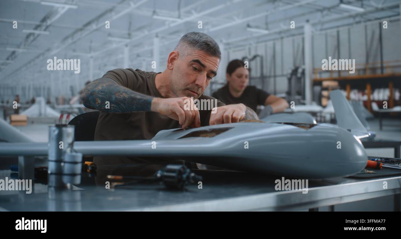 Specialista maschile esperto installa hardware elettronico, unità cilindrica e collega il cablaggio all'interno di un drone ad ala fissa, dimostrando l'assemblaggio preciso di UAV in una moderna struttura di produzione aerospaziale. Foto Stock