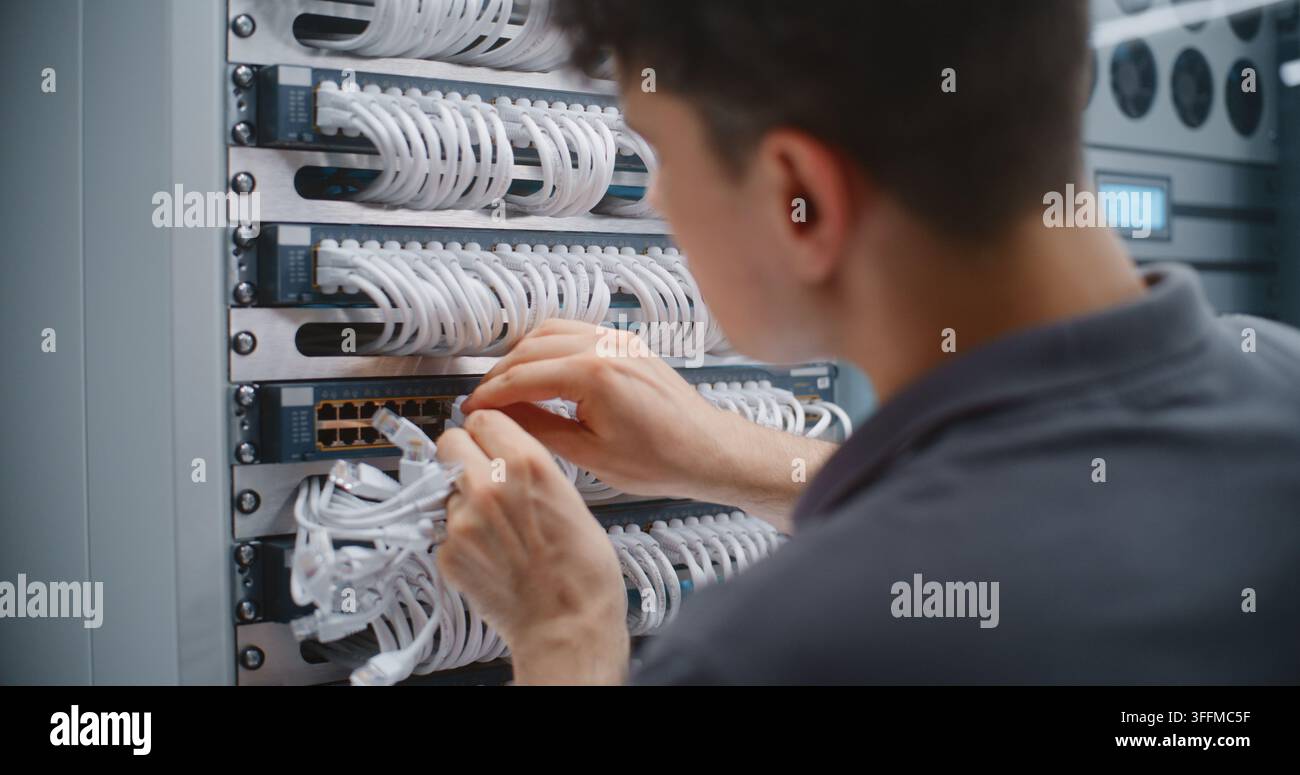 Tecnico di rete maschile esegue il patching del cavo a livello fisico all'interno del rack del server, regolando le connessioni Ethernet e i percorsi dei cavi per supportare un'infrastruttura organizzata e l'affidabilità della rete. Foto Stock