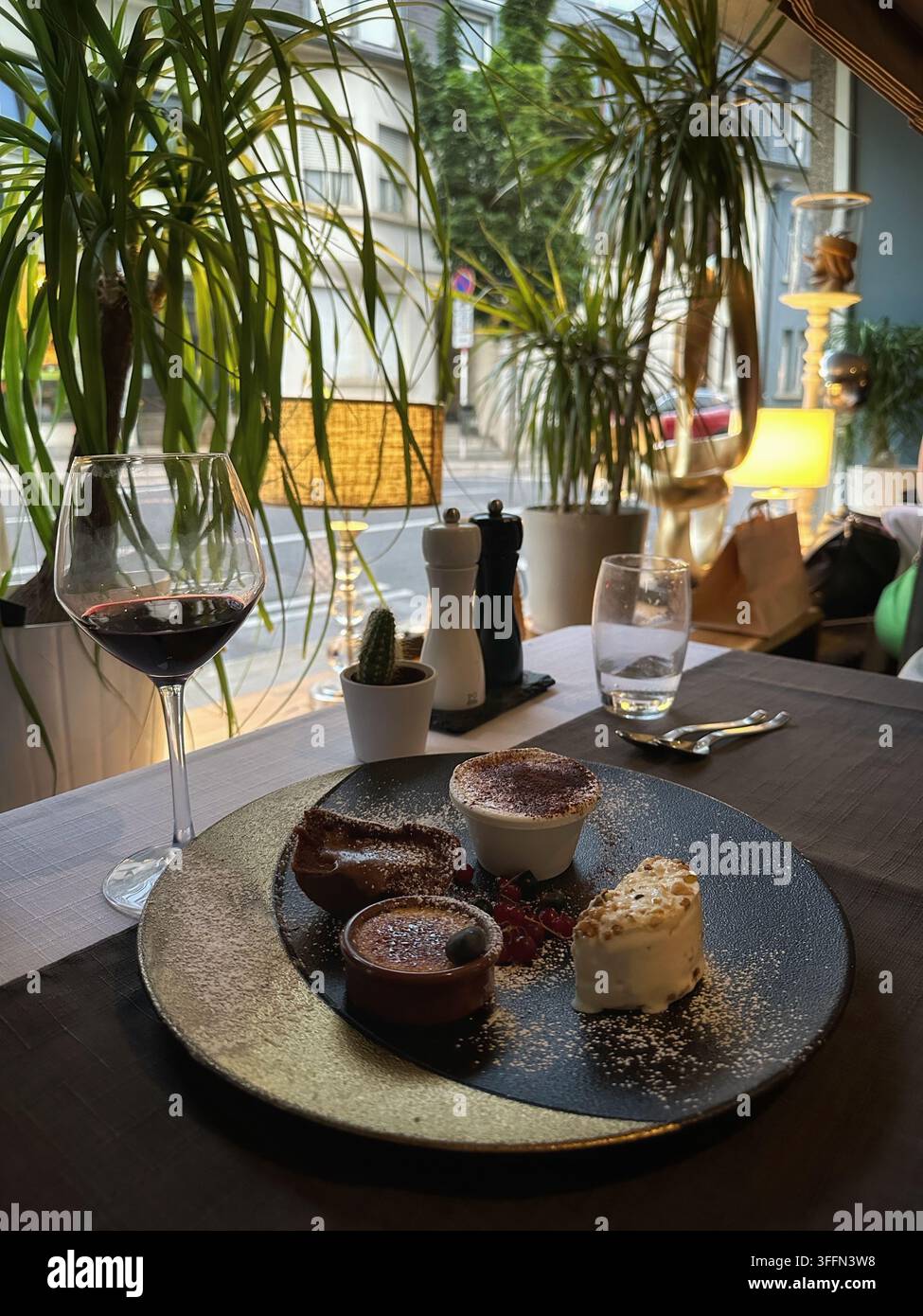 Piatto di dessert gourmet servito con un bicchiere di vino rosso in un interno di ristorante elegante. Un assortimento di dolci su un piatto scuro è accanto Foto Stock