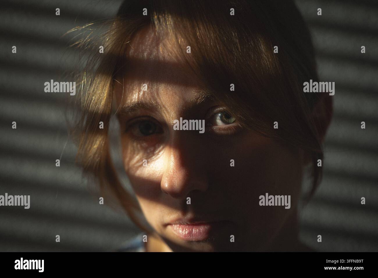 Ritratto ravvicinato di una giovane donna seria che guarda direttamente la fotocamera con luce solare geometrica e motivi di ombra delle luci sul viso. Foto Stock