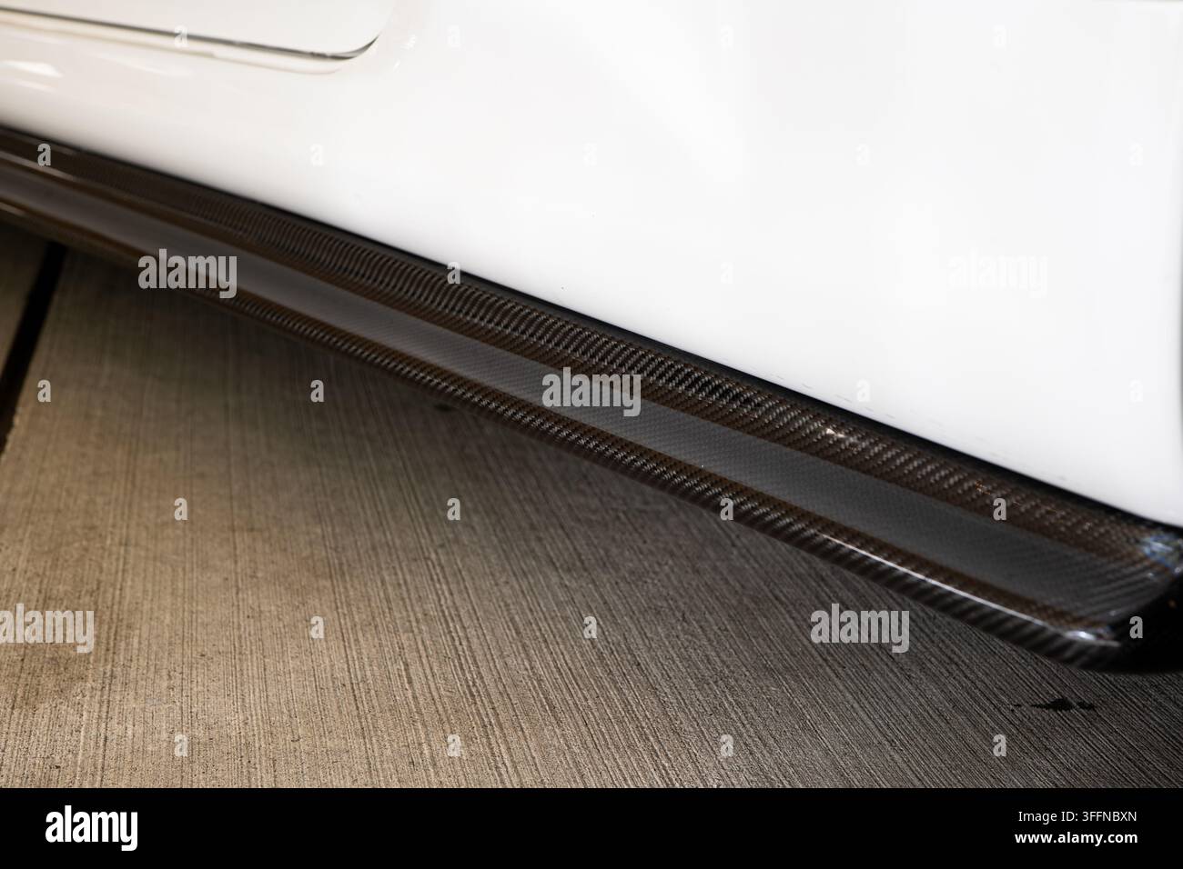 Vista ravvicinata ad angolazione bassa che mostra il motivo intrecciato di un'estensione del paraurti laterale in fibra di carbonio montata lungo il pannello inferiore bianco della carrozzeria di un'auto preparata. Foto Stock