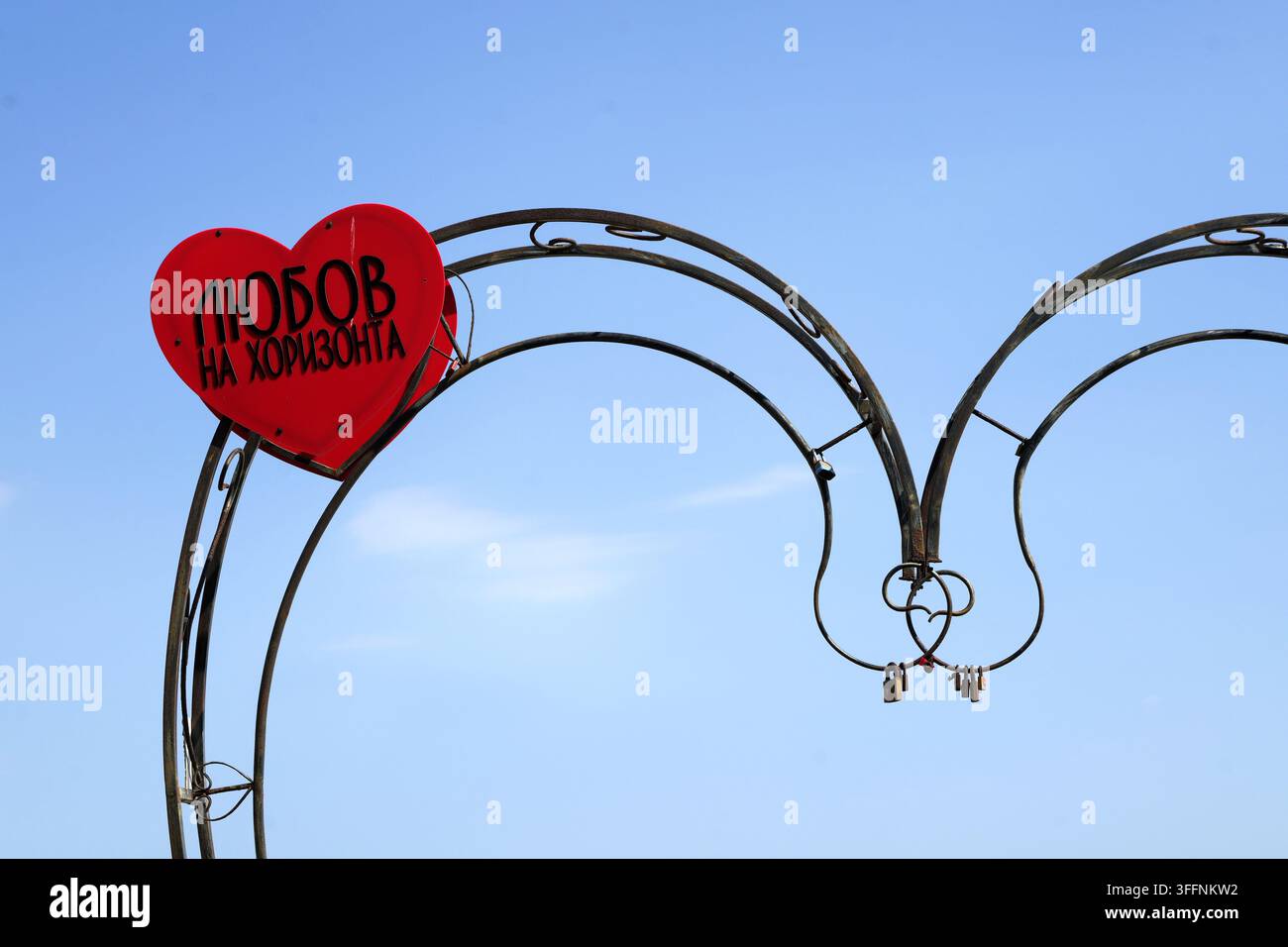 Segno a forma di cuore rosso che mostra l'amore all'orizzonte in testo bulgaro pende da un arco in ferro battuto adornato con lucchetti appesi. Simboleggia un amore duraturo Foto Stock