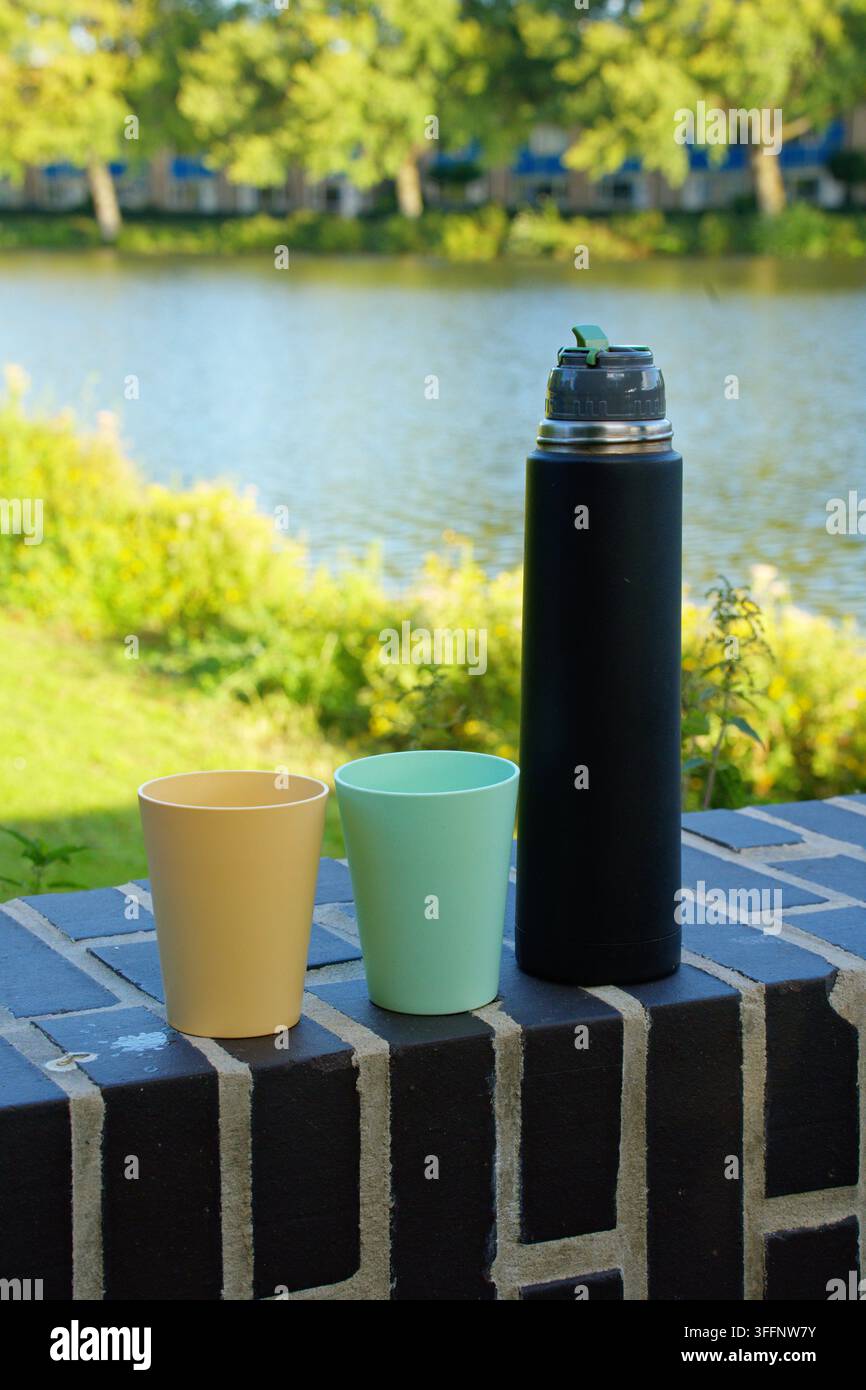 Un thermos nero e due bicchieri di plastica – uno giallo e uno verde menta – appoggiati su una superficie di mattoni vicino a un corpo d'acqua. Foto di alta qualità Foto Stock