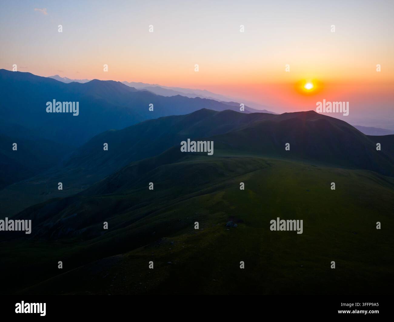 Una vista aerea cattura uno splendido paesaggio montano al tramonto in Kirghizistan. Dolci colline verdi e picchi stratificati creano un'atmosfera serena, arricchita da un cielo vivace. Foto Stock