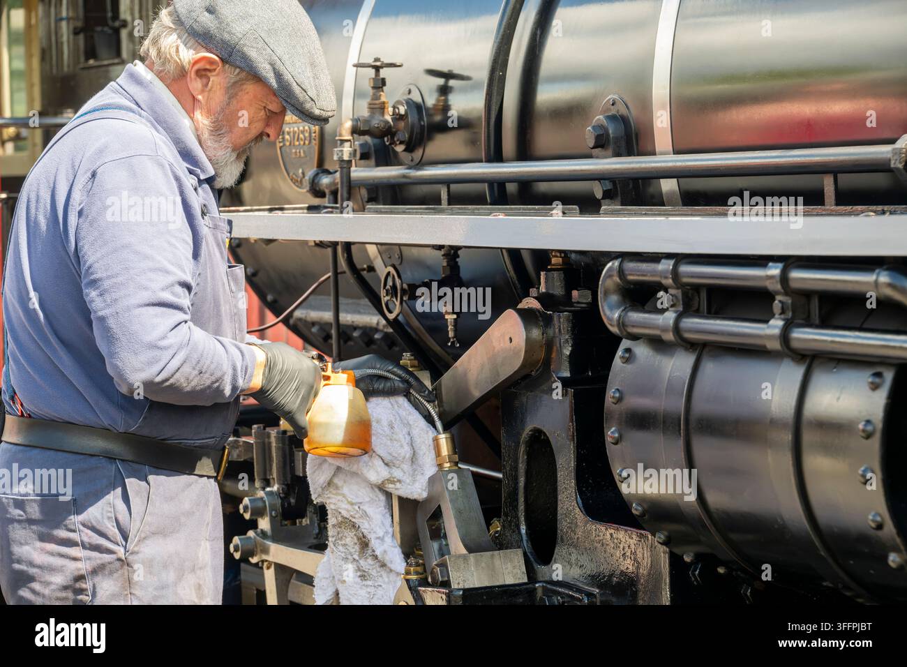Un uomo in tute e cappello piatto applica olio alla complessa meccanica di un treno a vapore storico, evidenziando manutenzione ed eredità Foto Stock