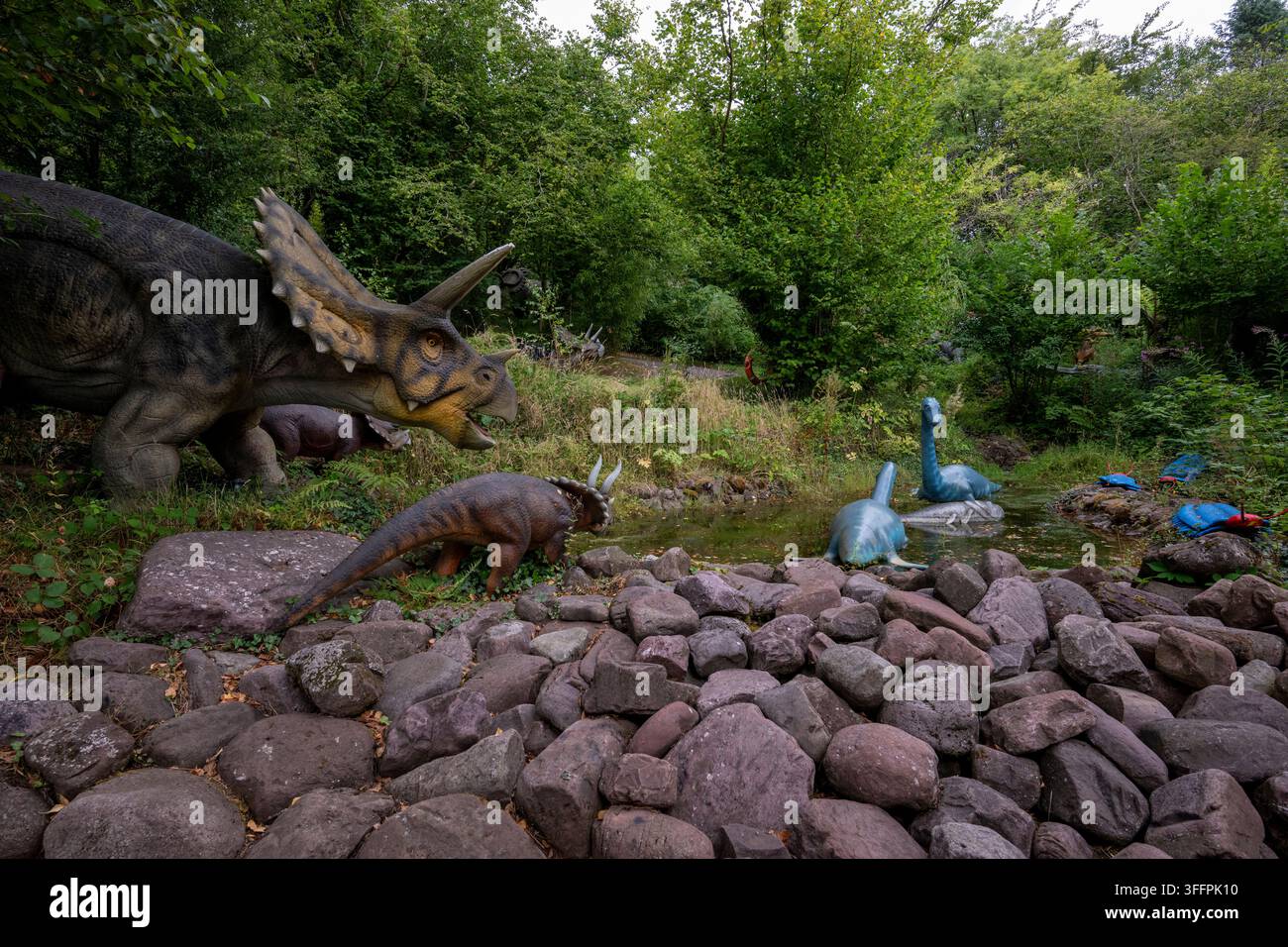 Sculture di dinosauri realistiche sono esposte in un ambiente naturalistico con una fitta vegetazione e una tranquilla fontana d'acqua Foto Stock