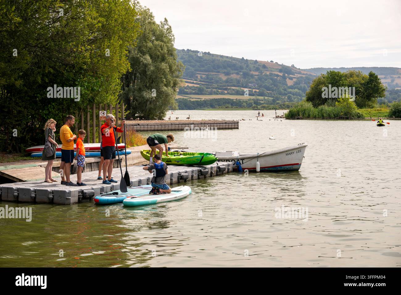 Un gruppo di persone si raduna su un molo galleggiante, pronto a fare paddleboard e kayak su un lago calmo circondato da rigogliose colline verdi e alberi Foto Stock