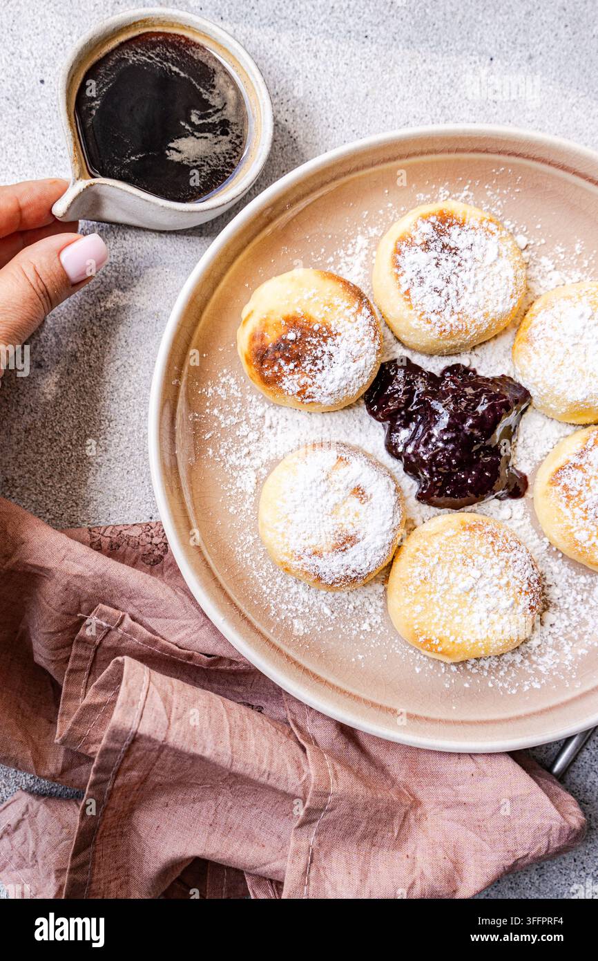 Vista dall'alto di syrniki ucraini tradizionali, pancake al formaggio spolverati di zucchero a velo, serviti con marmellata di mirtilli su un piatto di ceramica. Foto Stock