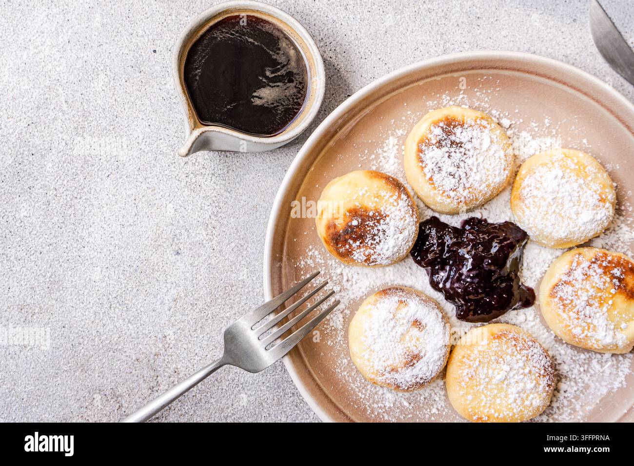 Vista dall'alto di pancake di formaggio syrniki fatti in casa spolverati di zucchero a velo, serviti con marmellata di mirtilli su un piatto di ceramica accanto a una tazza di caffè. Foto Stock