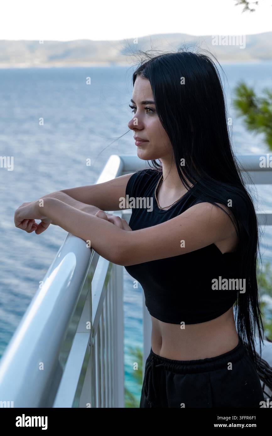 Una giovane donna in nero si trova su un balcone, guardando verso l'acqua. Foto Stock