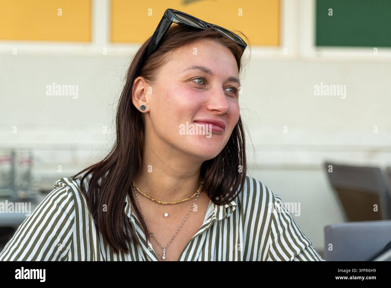 Una giovane donna sorridente è seduta all'aperto, indossa occhiali da sole sulla testa e una maglietta a righe. Foto Stock