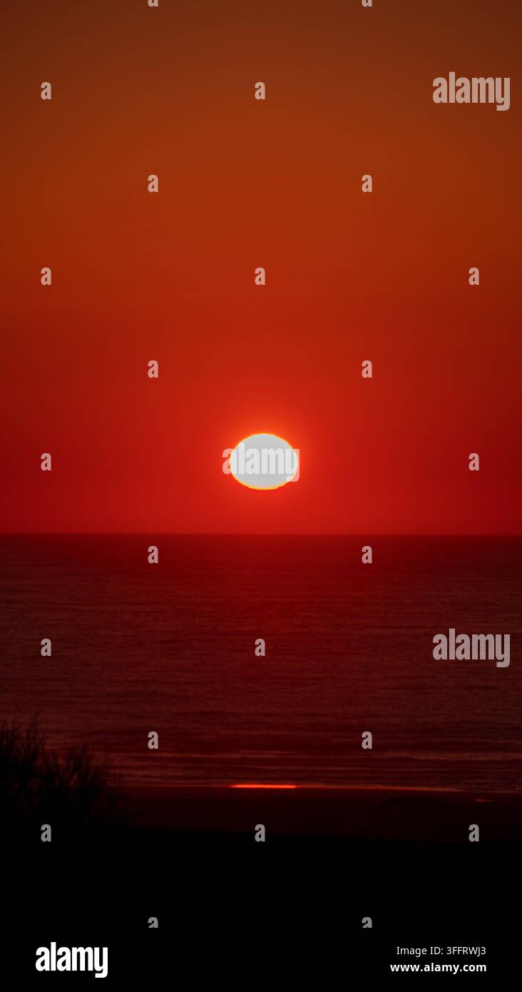 Scatto verticale del disco solare pieno che tramonta in un intenso cielo rosso al crepuscolo sopra l'orizzonte calmo dell'Oceano Atlantico in Marocco Foto Stock