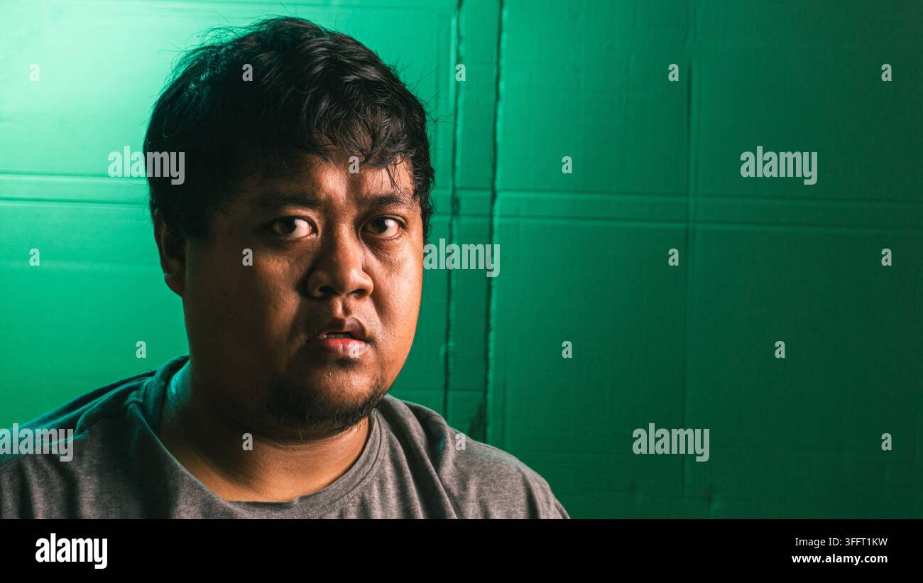 Un ritratto ravvicinato cattura un giovane indonesiano con un'espressione facciale sbalordita e vuota su uno sfondo verde vivace e texture. Il Foto Stock