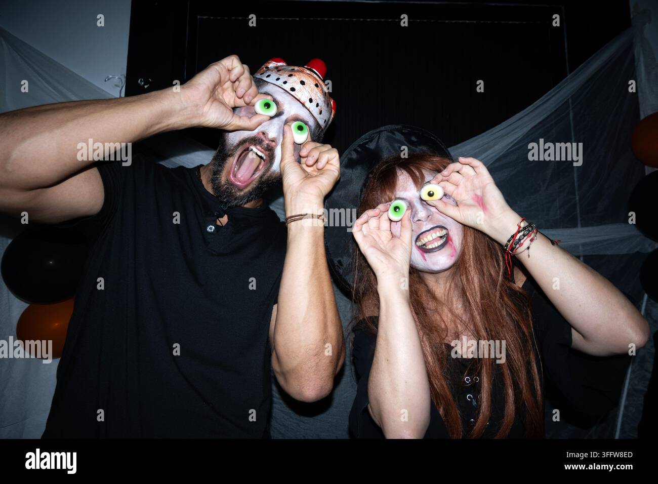 Uomo e donna in costumi di Halloween che tengono occhi di caramelle sopra i loro occhi e fanno espressioni divertenti a una festa. Foto Stock