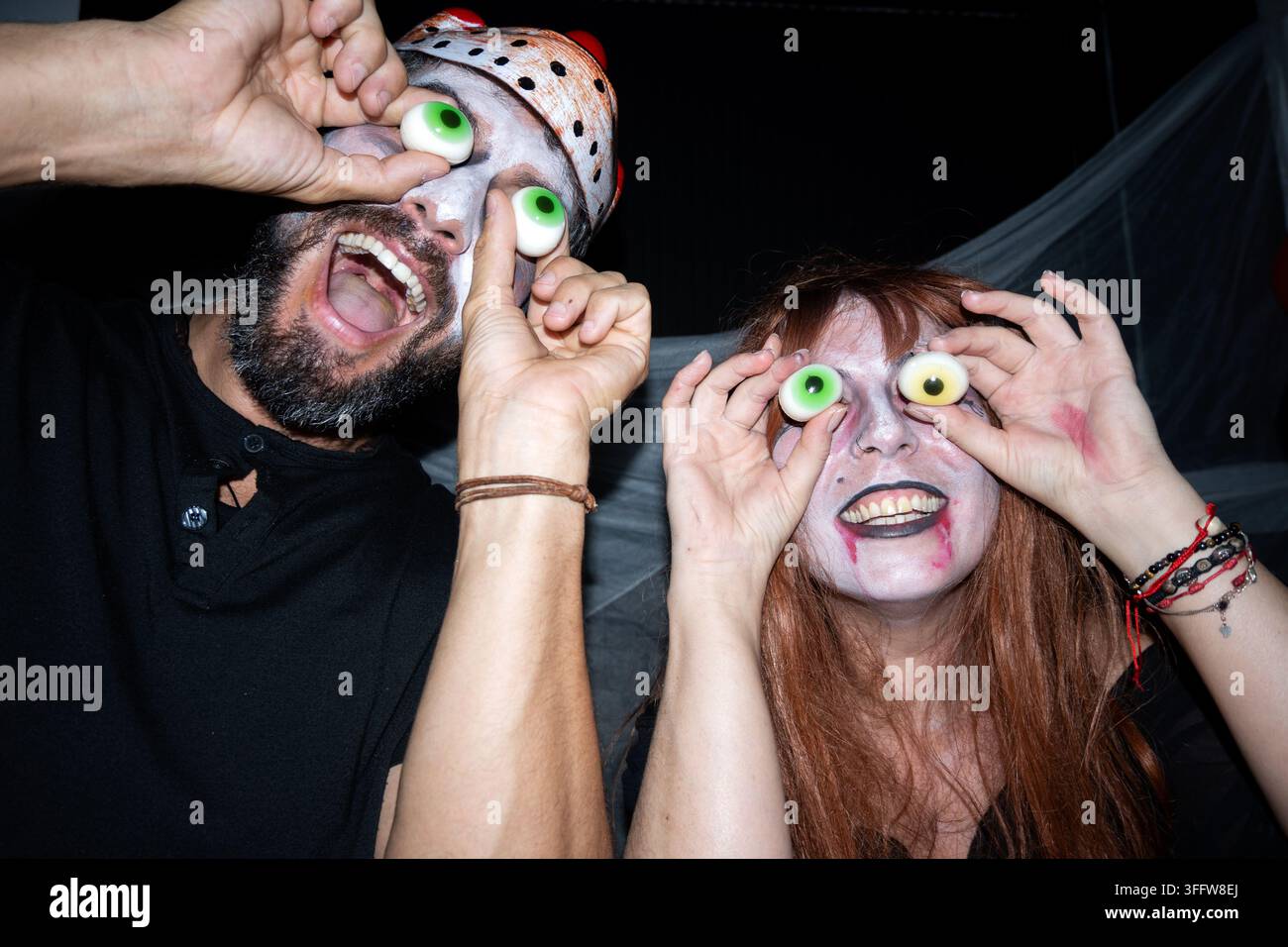 Uomo e donna in costumi di Halloween che tengono occhi di caramelle sopra i loro occhi e fanno espressioni divertenti a una festa. Foto Stock