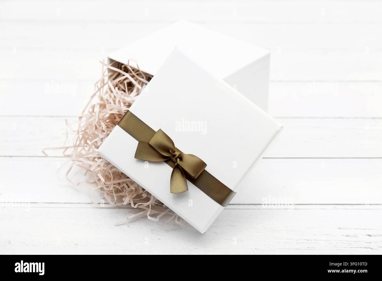 Elegante scatola regalo bianca aperta con fiocco di nastro dorato e imbottitura di carta strappata beige su uno sfondo di legno bianco, perfetta per confezioni di lusso. Foto Stock