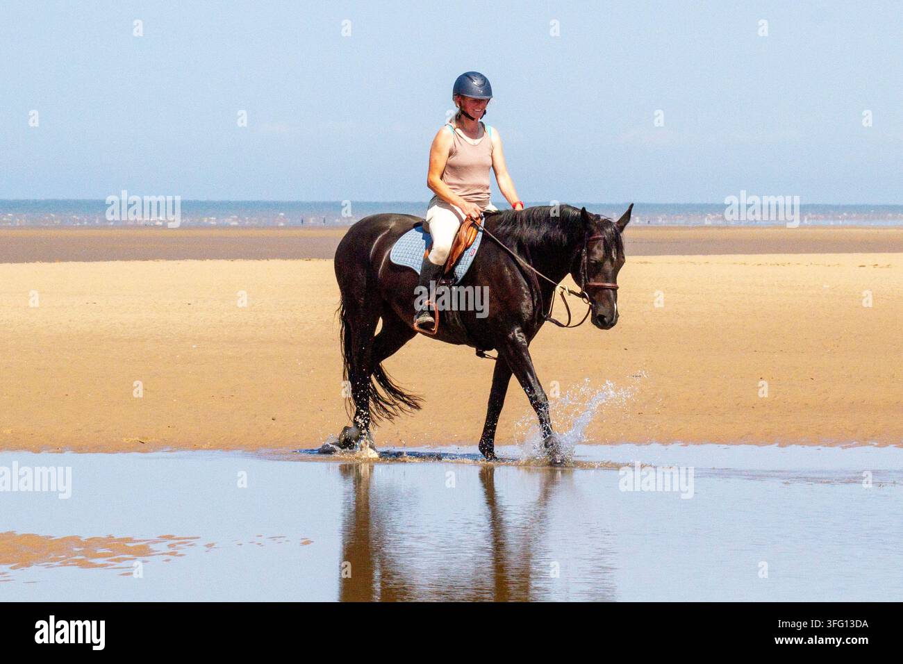 Una cavallerizza sorridente guida un potente cavallo sportivo irlandese nero attraverso una pozza di marea poco profonda su una vasta spiaggia sabbiosa. Southport, Regno Unito. Foto Stock