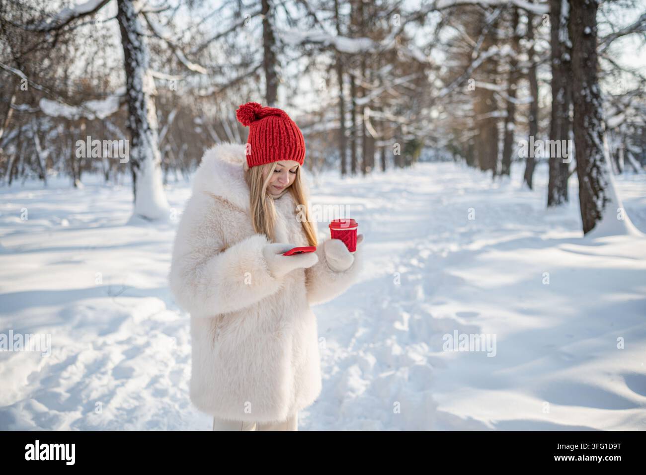 Giovane donna controlla lo smartphone mentre tiene una bevanda calda da asporto durante una passeggiata invernale, combinando comunicazione, calore e tempo libero accogliente per le vacanze. Foto Stock