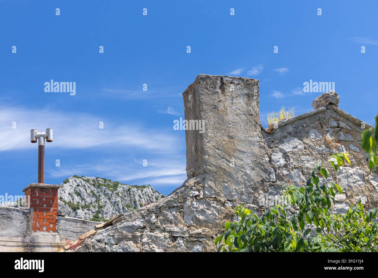 Tetto di pietra consumato dal tempo e camino che si ergono sotto un cielo estivo blu brillante, con creste rocciose e fogliame verde in primo piano che trasmettono Foto Stock