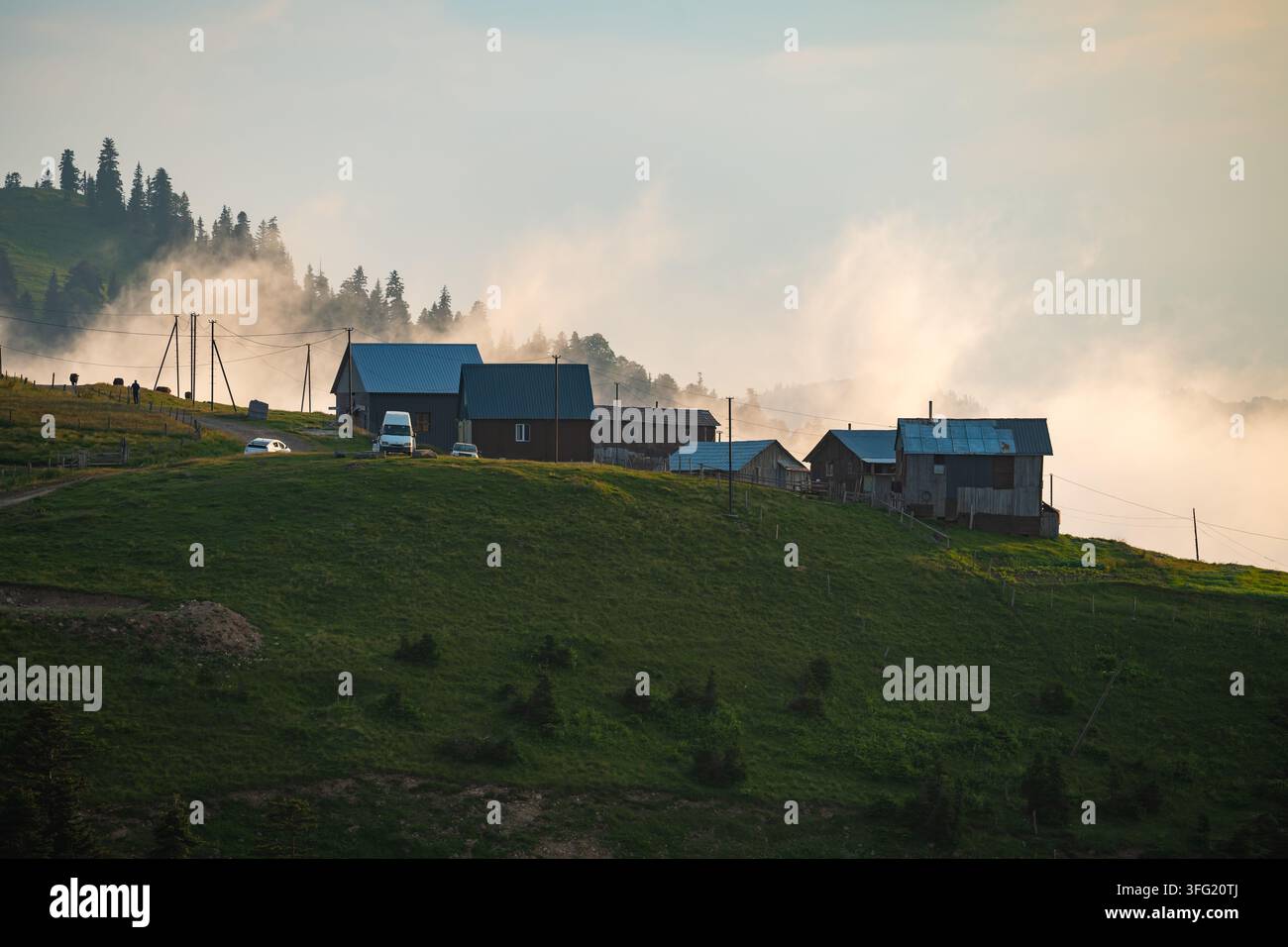Monti Adjara, Georgia. Un gruppo di case di legno rustiche si trova su una collina verdeggiante avvolta da una nebbia mattutina eterea, con lontani boschi di pini vi Foto Stock
