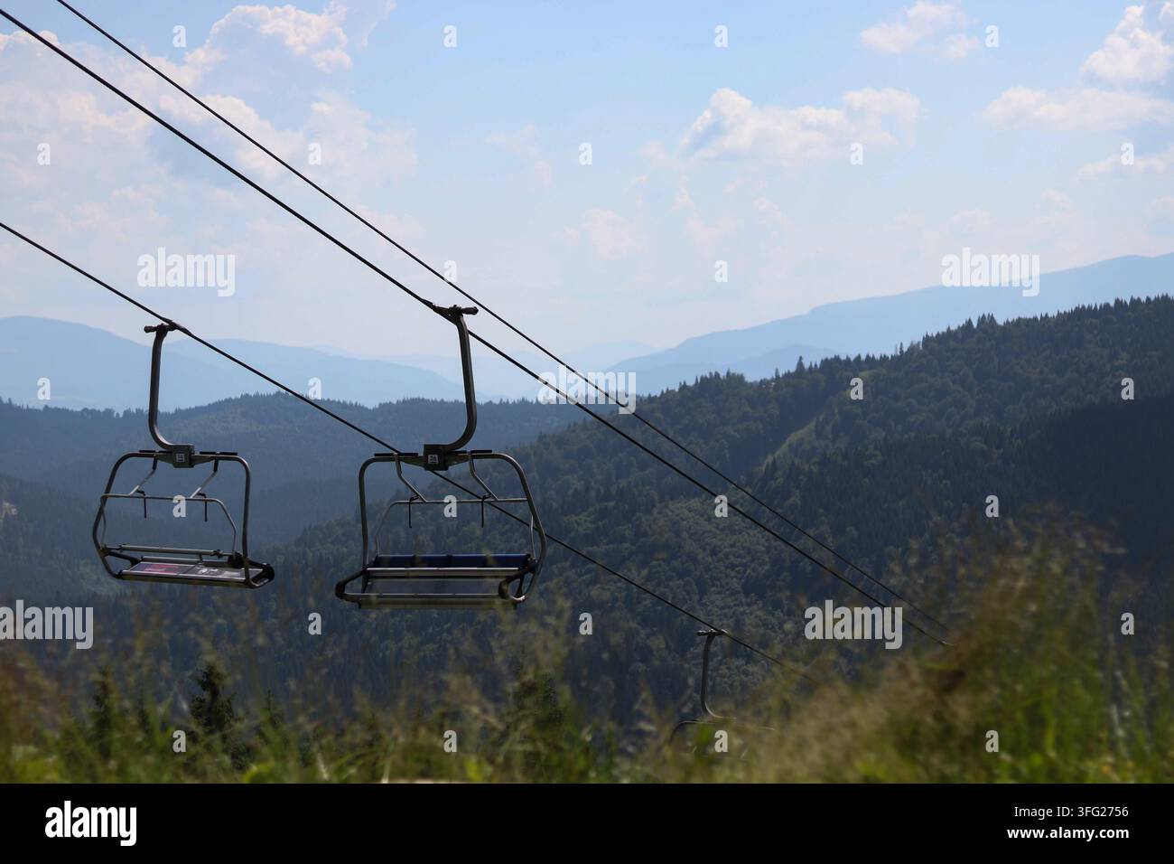 I sedili vuoti della seggiovia pendono sopra i pendii verdi delle montagne sotto un cielo soleggiato. Una scena tranquilla che simboleggia il turismo estivo e la libertà dei viaggi in montagna. Foto Stock