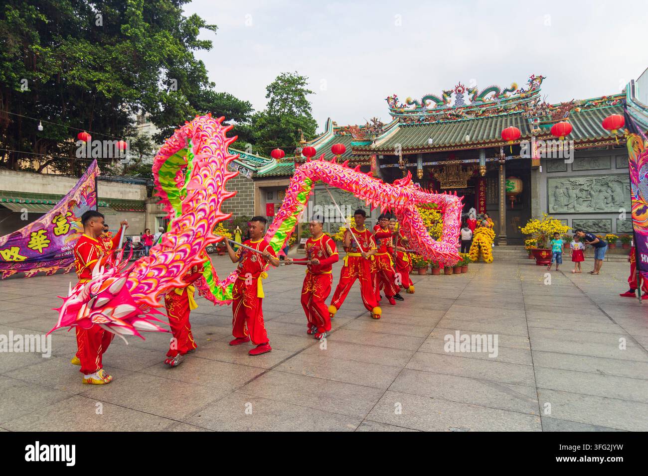 Una compagnia in costumi rossi esegue una colorata danza del drago davanti a un tempio ornato mentre gli spettatori osservano dal cortile. Foto Stock