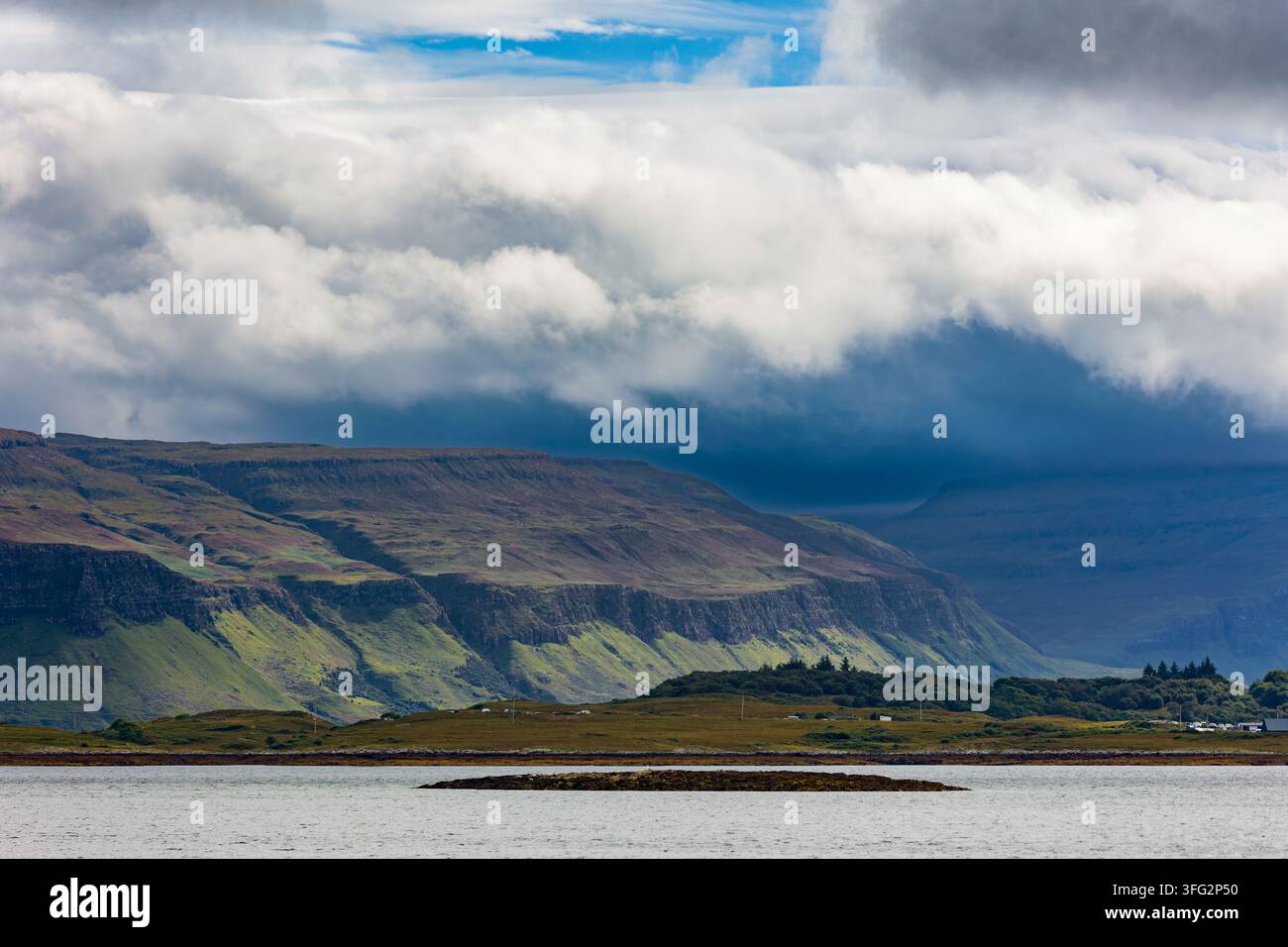 Strato drammatico di nuvole che proietta ombre su pendici montuose terrazzate e costa erbosa, baia tranquilla sotto scogliere di altopiano frastagliate Foto Stock