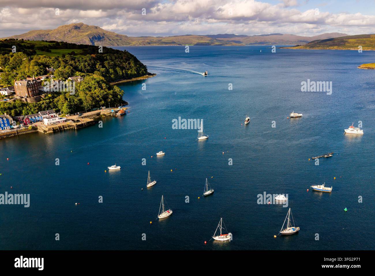 Baia di Tobermory sull'Isola di Mull, yacht all'ancora in un braccio di mare calmo con costa boschiva, molo e montagne sotto nuvole sparse nella Scozia occidentale Foto Stock
