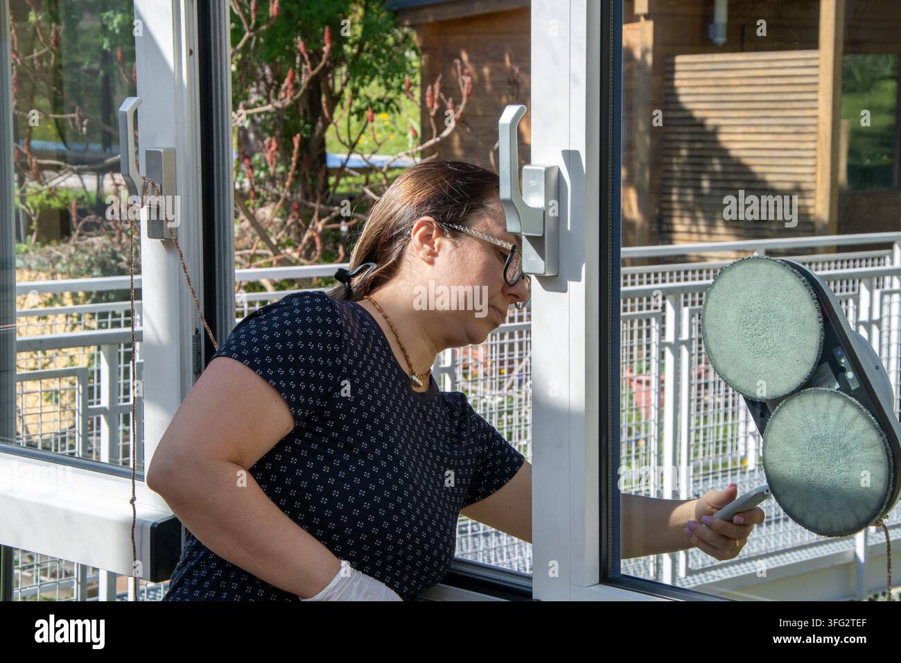 Una donna pulisce una finestra di vetro usando un lavavetri robotico in una giornata soleggiata. Foto Stock