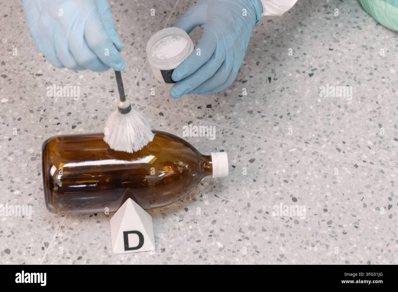 Un investigatore forense guantato applica polvere per impronte digitali con un pennello su una bottiglia ambrata accanto al marcatore delle prove D durante l'esame della scena del crimine. Foto Stock