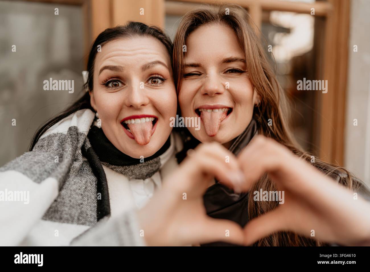 Selfie tra amici a forma di cuore con due allegre giovani donne che tirano fuori la lingua e formano una forma a cuore all'aperto durante l'autunno Foto Stock