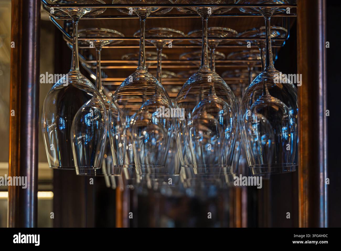 Calici appesi a una rastrelliera in un interno di bar caldo. File di calici vuoti appesi a testa in giù da una rastrelliera metallica, riflettendo luce calda e fredda Foto Stock