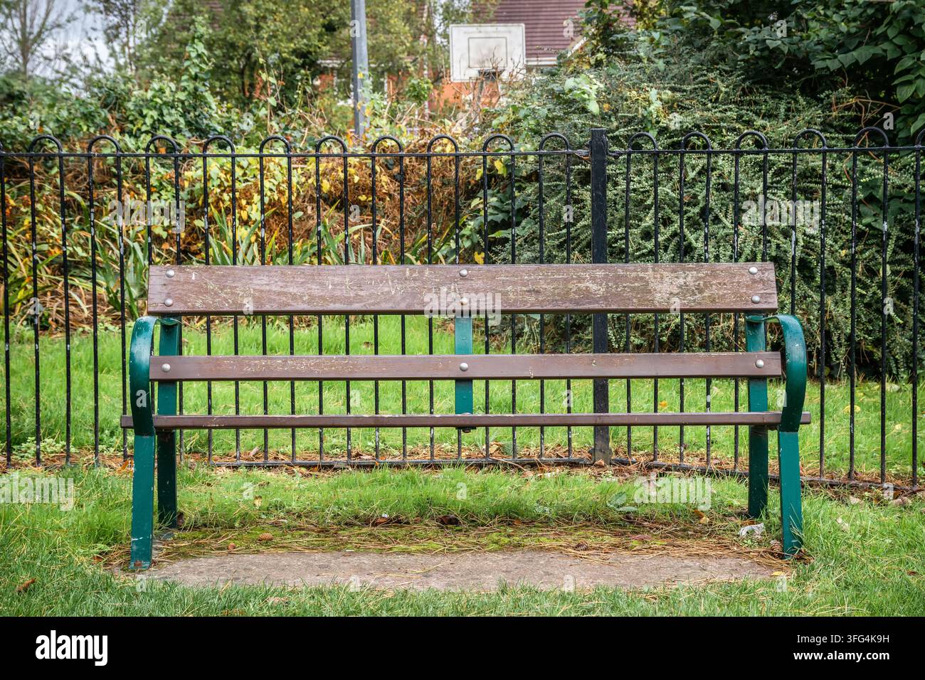 Una panchina vuota in doghe di legno con braccioli in ferro verde posizionati simmetricamente davanti a ringhiere metalliche nere in un giardino pubblico urbano. Foto Stock