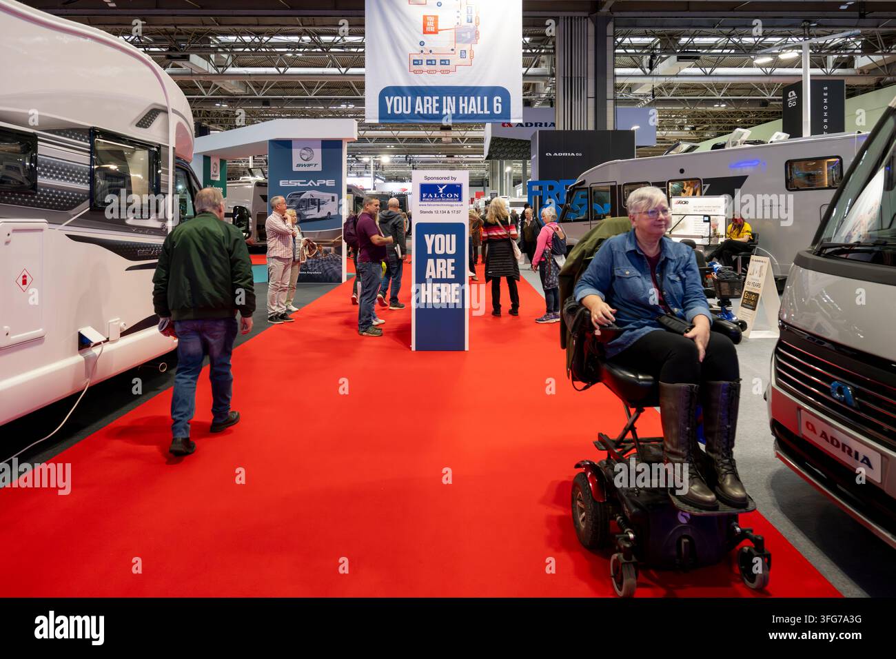 I visitatori esplorano camper e roulotte a una fiera, alcuni con ausili per la mobilità sul vivace pavimento rosso Foto Stock
