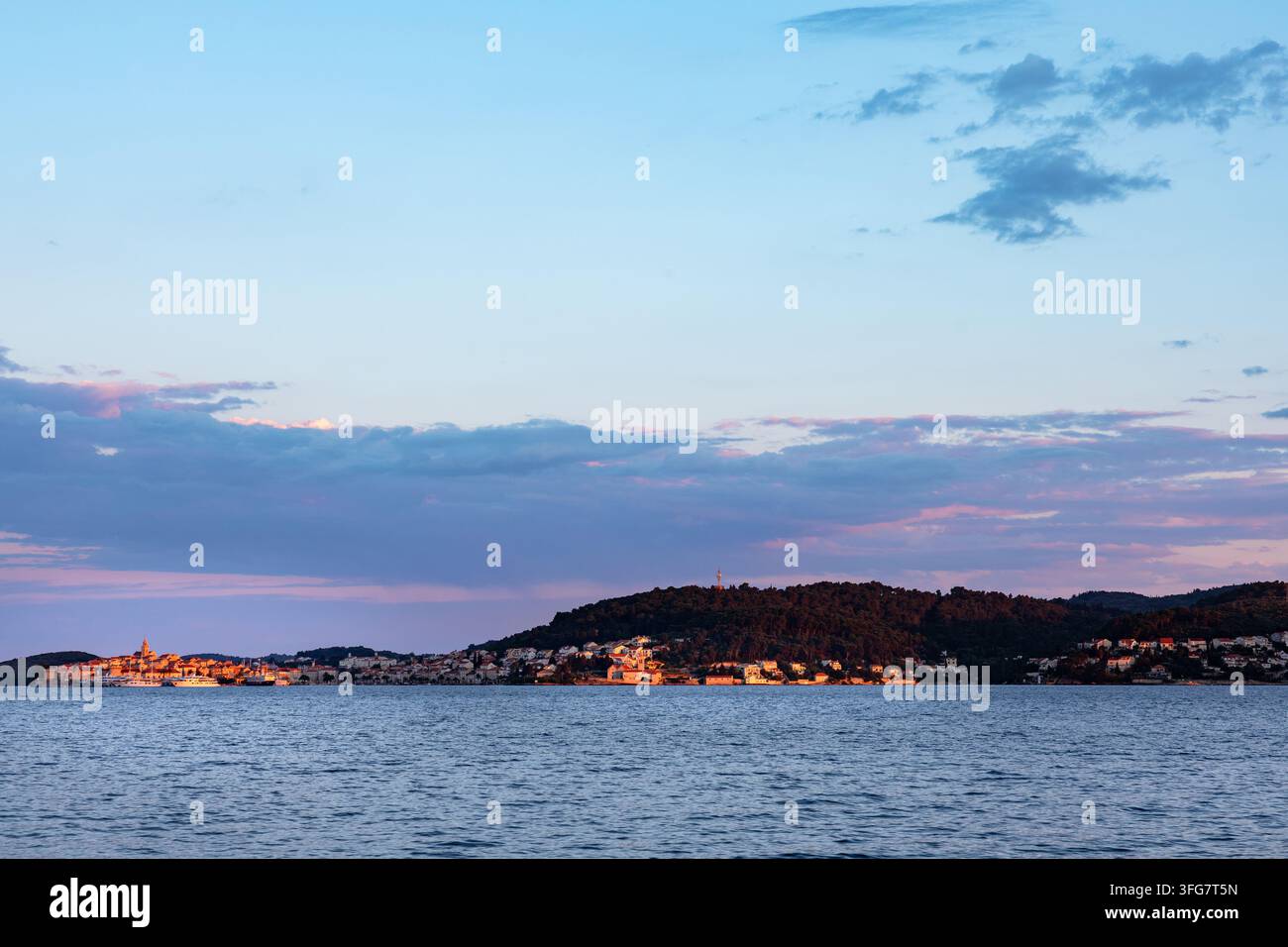 Una tranquilla città costiera immersa nella calda luce dorata dell'ora blu, incorniciata da dolci colline, nuvole drammatiche e acque marine calme al tramonto. Atmosfera serena id Foto Stock