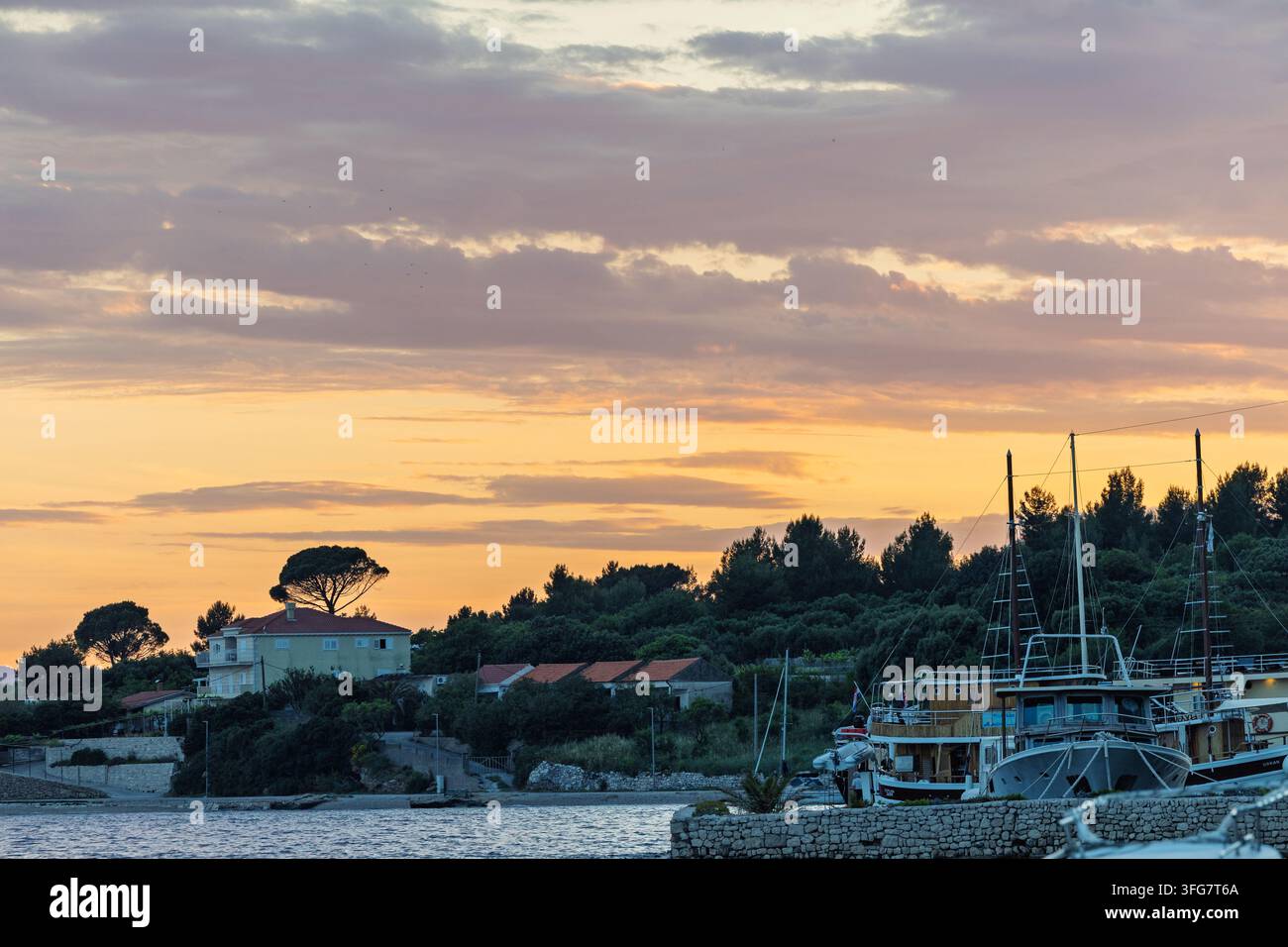 Il cielo dorato del tramonto proietta una luce calda su un tranquillo porto costiero con barche ormeggiate, molo in pietra e villaggio alberato sulla riva, evocando calma, tra Foto Stock