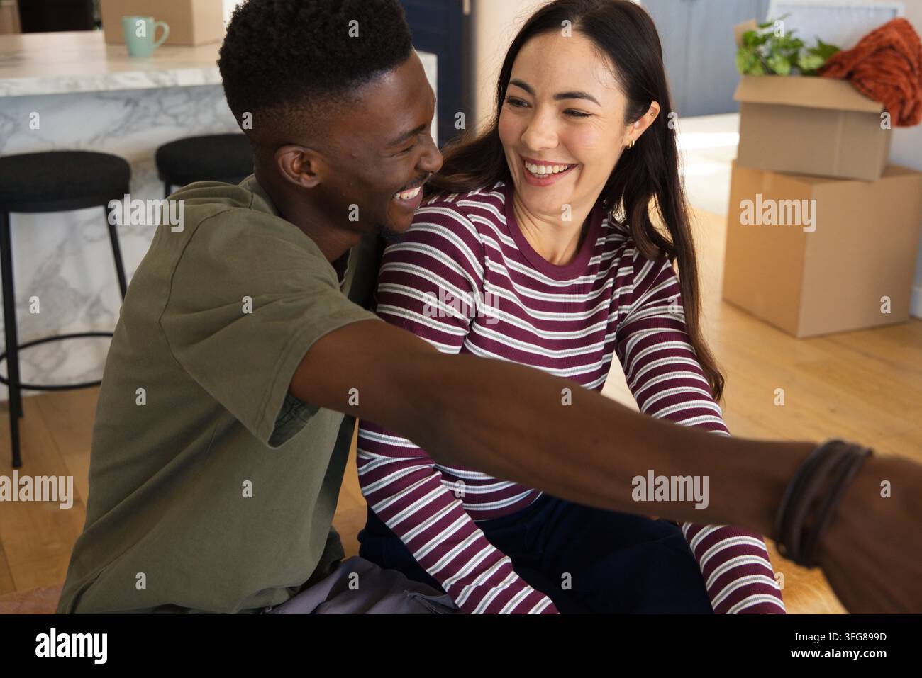Coppia seduta sul pavimento in legno duro della cucina, sorridente e appoggiata vicino a scatole da trasloco e bancone di marmo Foto Stock