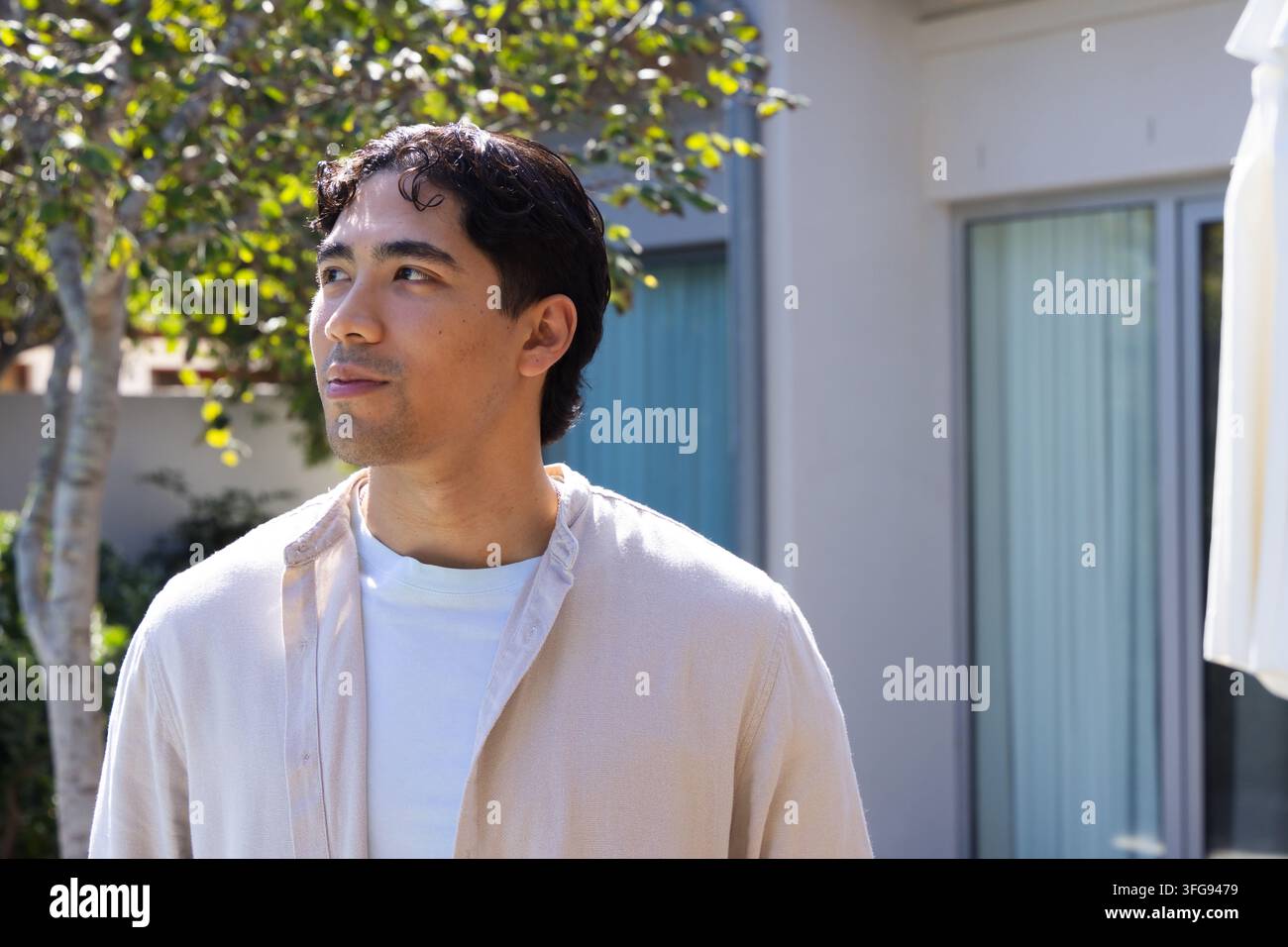 Uomo asiatico adulto in piedi sul patio nella luce screziata, che guarda a sinistra verso un albero, indossa una camicia chiara Foto Stock