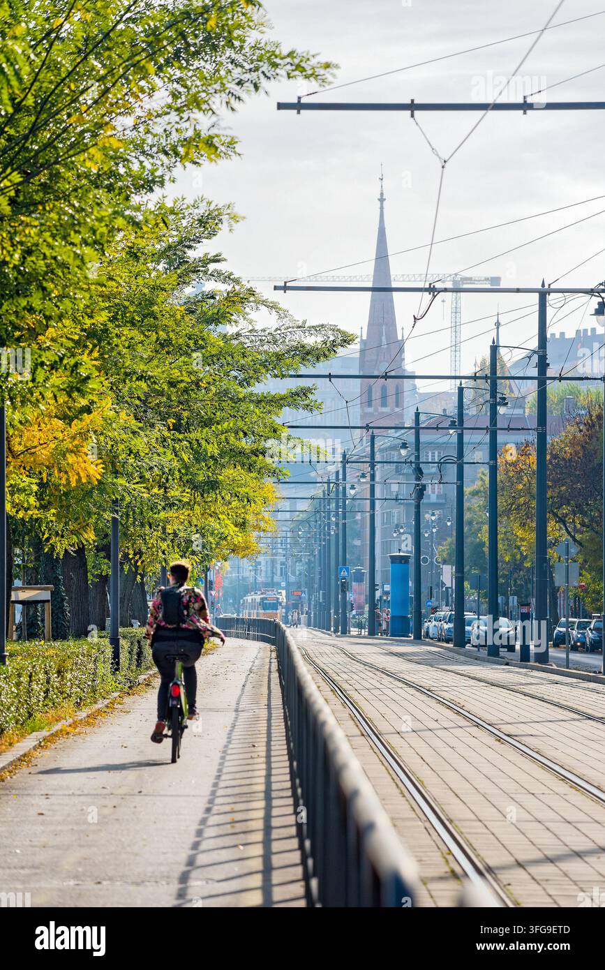 Ciclista che pedala lungo una pista ciclabile urbana accanto ai binari del tram a Budapest in una giornata autunnale soleggiata, con una guglia di chiesa in lontananza Foto Stock