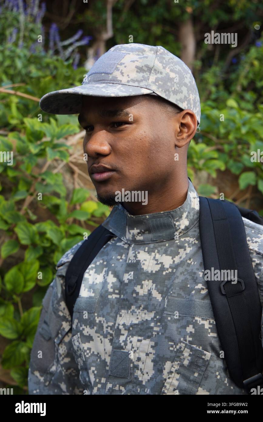 Uomo adulto africano con cappello e giacca mimetica digitale in piedi in un giardino frondoso con uno zaino Foto Stock
