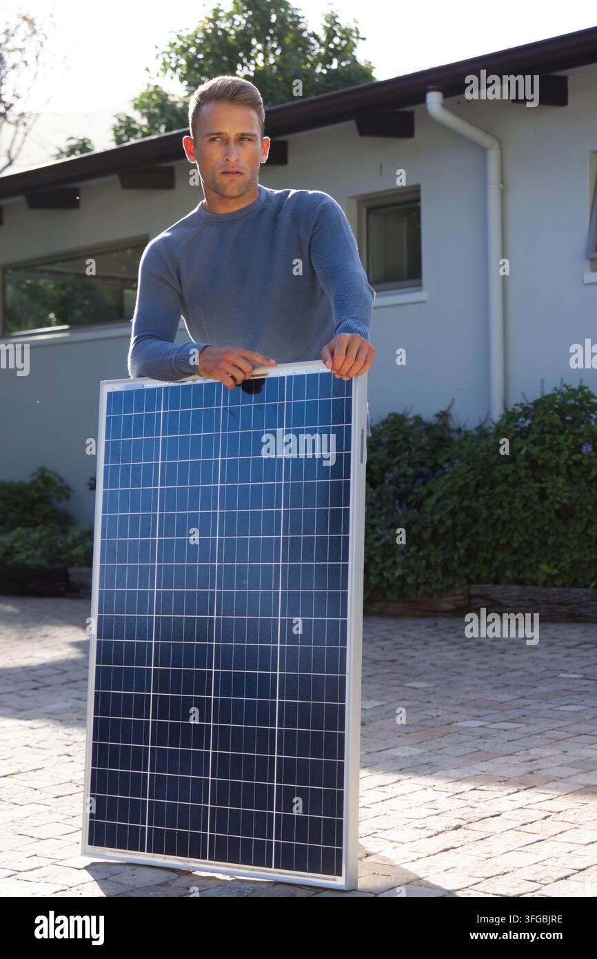 Uomo che tiene un pannello solare in posizione verticale nel vialetto di una casa a un piano, indossa un maglione grigio Foto Stock