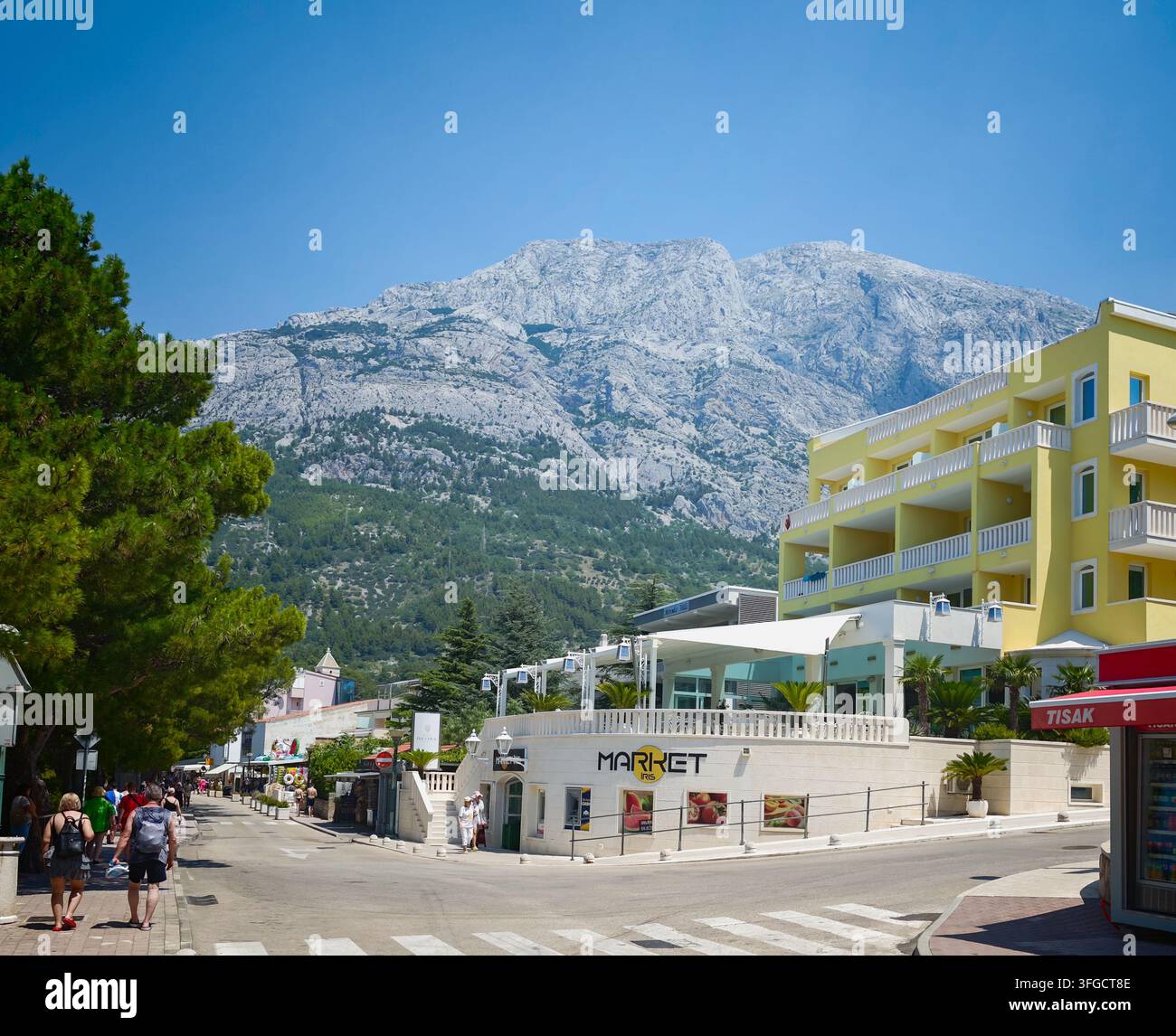 I turisti passeggiano per una strada soleggiata a Baska Voda sotto gli imponenti picchi calcarei del monte Biokovo sotto un cielo blu limpido. - Immagine stock catturata con smartphone