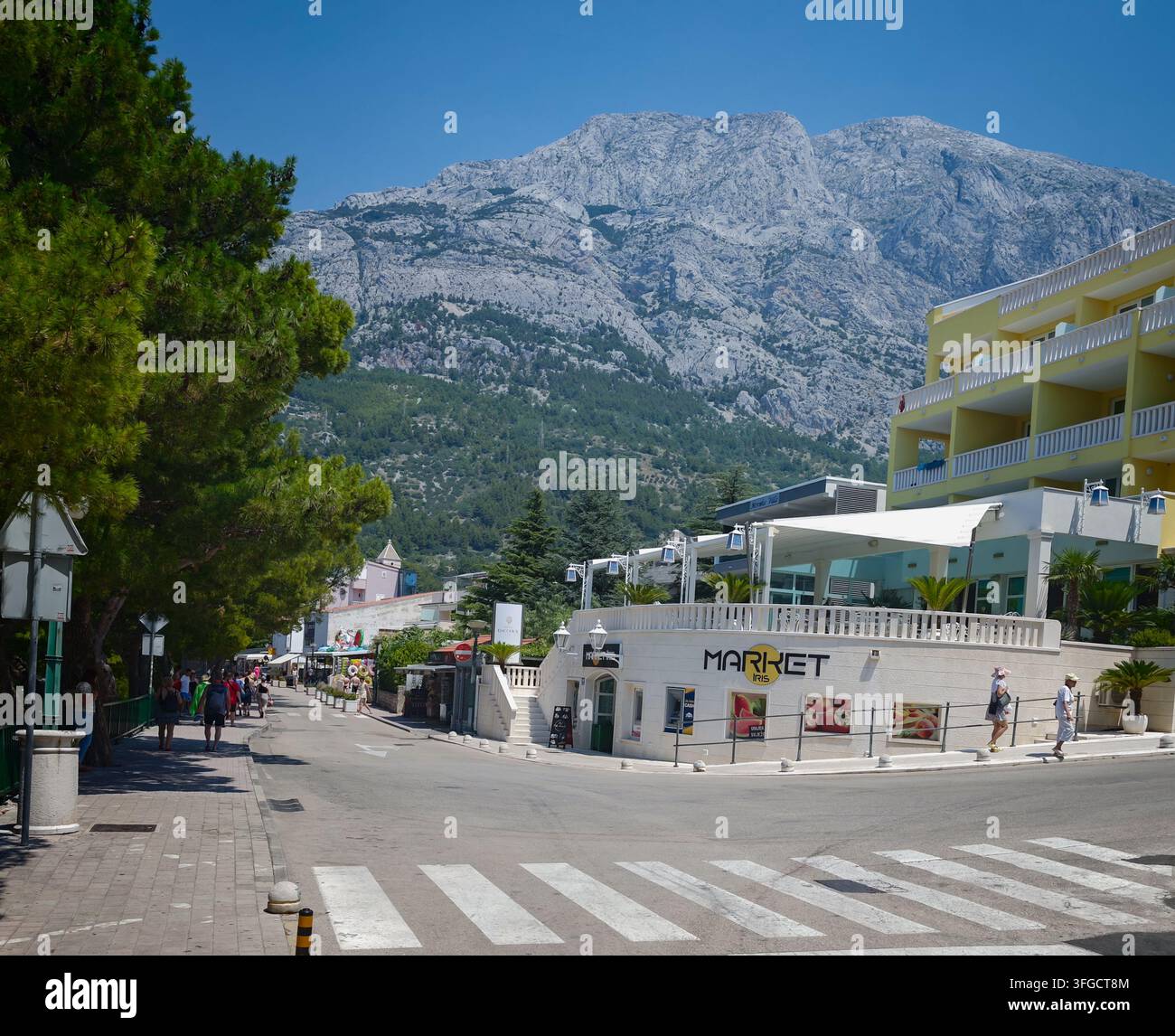 I turisti passeggiano per una strada soleggiata a Baska Voda sotto gli imponenti picchi calcarei del monte Biokovo sotto un cielo blu limpido. - Immagine stock catturata con smartphone