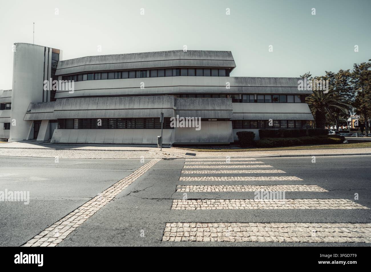Edificio modernista consumato con lunghe finestre orizzontali e forme angolari in cemento si affaccia su una strada vuota, dove strisce di attraversamento in ciottoli chiari conducono Foto Stock