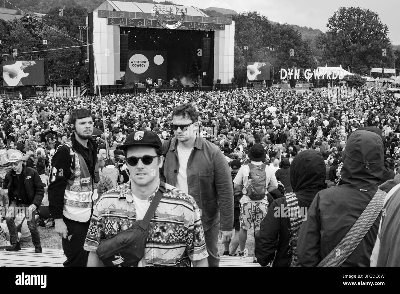 FOLLE DEL FESTIVAL FREDDA IN BIANCO E NERO, REPORTAGE, GREEN MAN FESTIVAL 2026, BRECON, GALLES, REGNO UNITO, 2026: In una fotografia in bianco e nero, diversi giovani uomini camminano tra la folla del festival, uno in primo piano indossa una camicia a motivi, occhiali da sole e un cappello. Foto: Rob Watkins. INFO: Il Green Man Festival si tiene nei Brecon Beacons ad agosto. Celebra musica alternativa, arti, letteratura, scienza e cultura familiare in uno splendido scenario montano. L’evento del 2026 si è svolto durante temporali alla fine di una lunga ondata di caldo e di siccità. Foto Stock