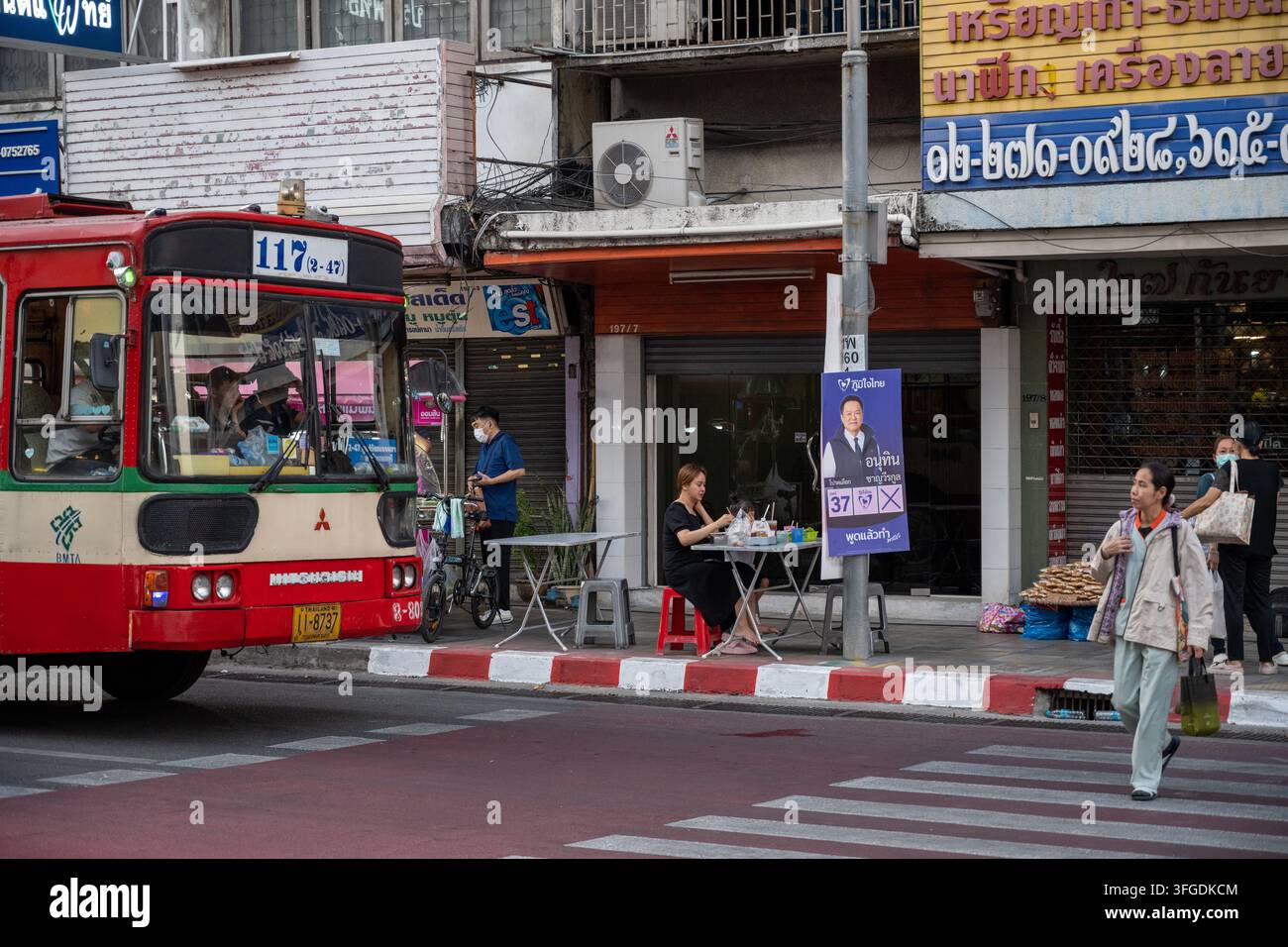 Chatumit Village, Bangkok, Thailandia, 3 febbraio 2026. Un autobus della linea 117 della BMTA passa davanti a pedoni e attività gastronomiche sul marciapiede in una strada commerciale. Foto Stock